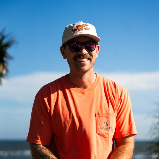 Man wearing an orange shirt and white cap with a logo, standing outdoors with a clear blue sky.