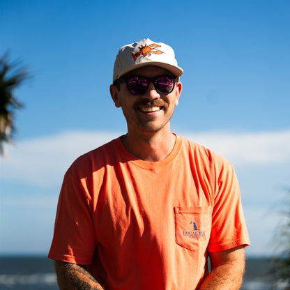 Man wearing an orange shirt and white cap with a logo, standing outdoors with a clear blue sky.