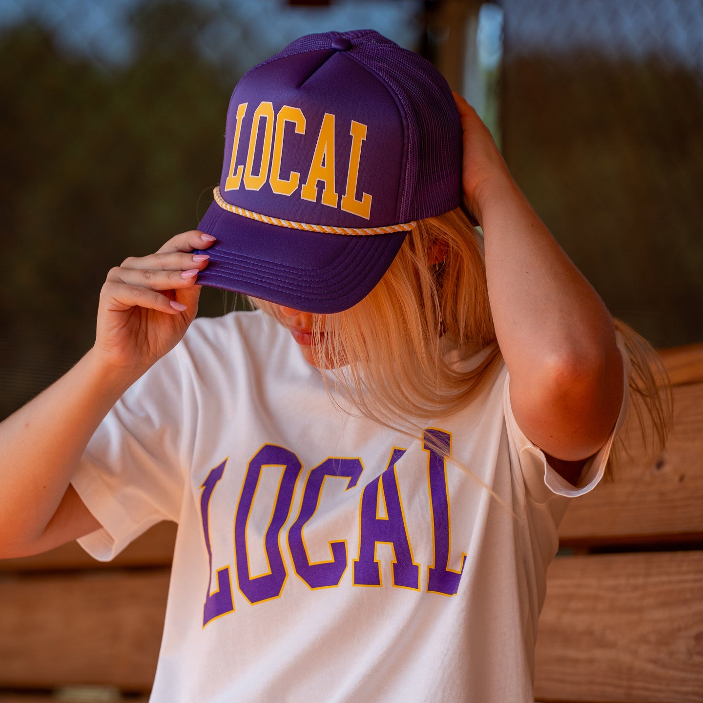 A woman wearing a purple and yellow trucker hat with 'LOCAL' printed on it, paired with a matching white t-shirt. She is holding the brim of the hat with one hand and adjusting it with the other, set against a warm, blurred outdoor background.