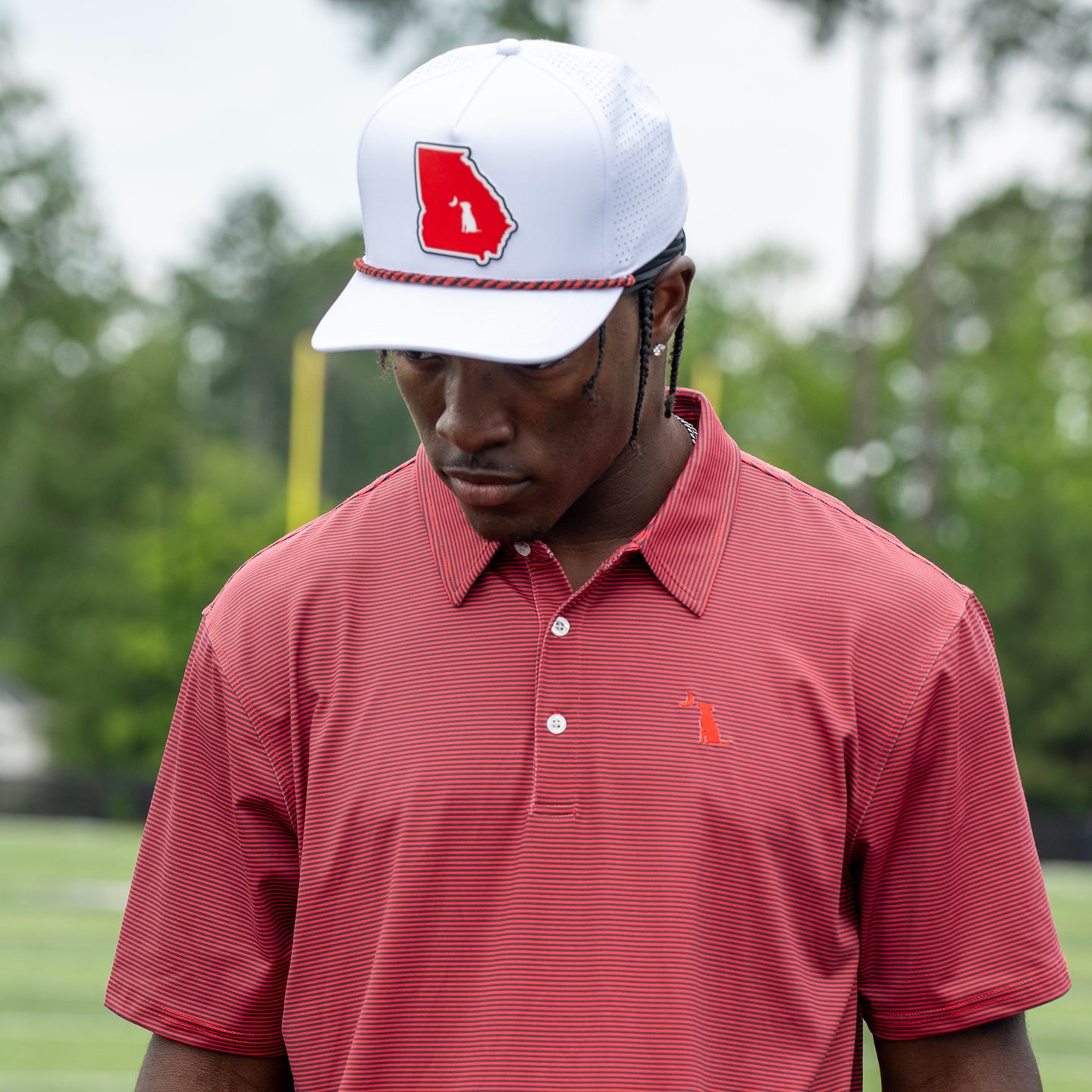 A man wearing a white baseball cap with a red Georgia logo and a red striped polo shirt, standing outdoors on a golf course.