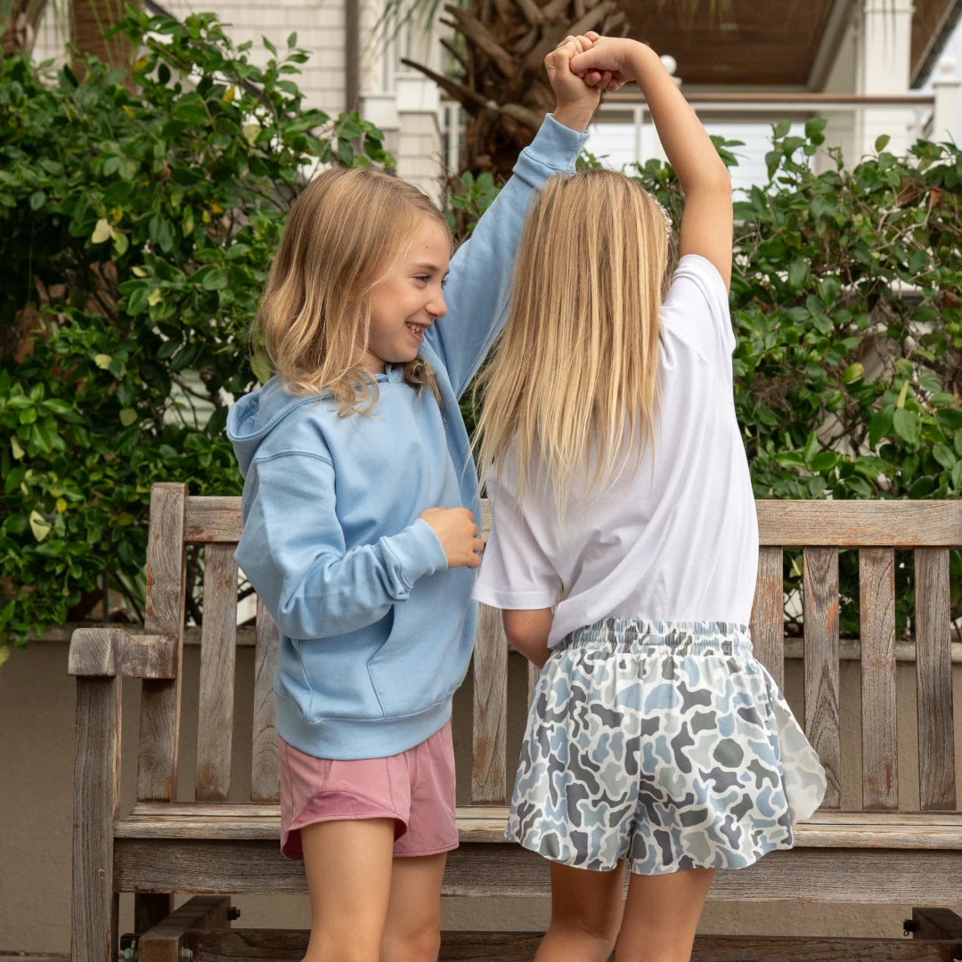 Two young girls standing on a wooden bench outdoors, holding hands. One girl is shown from the back wearing a white t-shirt and a gray patterned skirt, while the other is shown from the side/back wearing a light blue hoodie and pink shorts.