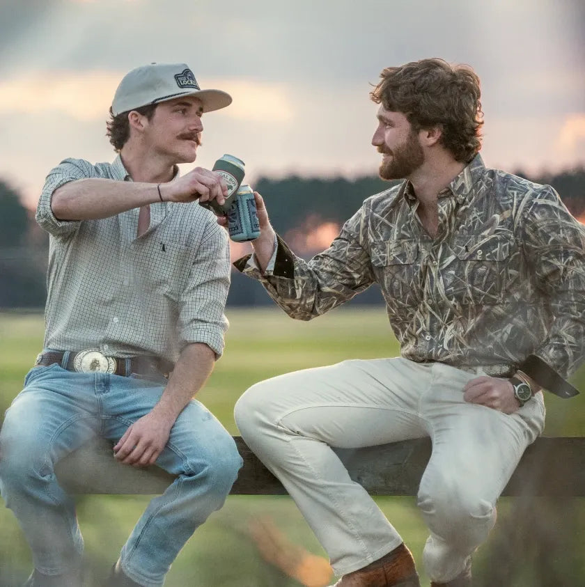 Two men sitting outdoors at sunset, each holding a green insulated mug. One wears a light gray button-up shirt and jeans, and the other wears a camouflage long-sleeve shirt and off-white pants.