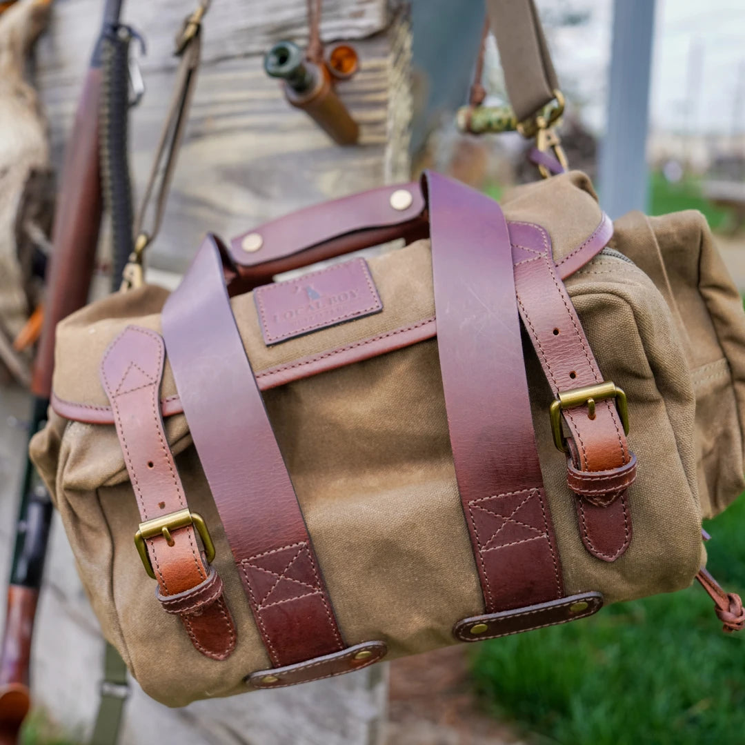 A canvas duffel bag with wide brown leather straps and brass buckles, shown hanging outdoors against a blurred background of gear and greenery.